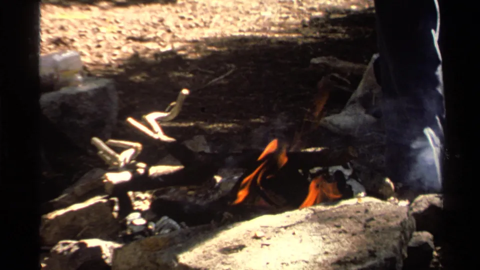 1978 Lake Tahoe home-movie frame of a small forest campfire with stones, smoke, and a standing adult partly visible nearby.