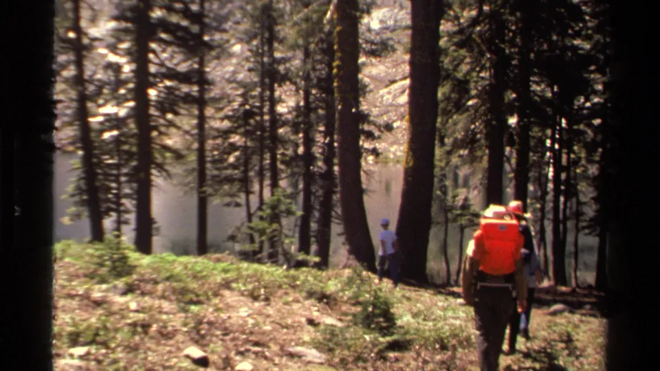 1978 Lake Tahoe home-movie still with a hiker carrying a bright orange backpack on a wooded slope beside visible water.