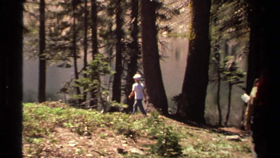 1978 Lake Tahoe frame showing a hiker in a white shirt and cap walking through pines with water visible behind the trees.