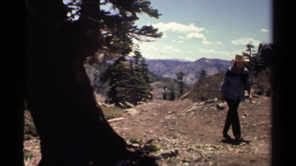 1978 Lake Tahoe-area 8mm frame showing a hiker on a rugged trail, with pine trees, rocky ground, distant mountains, and partly cloudy sky.