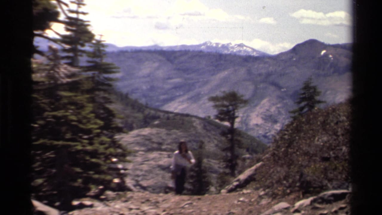 1978 Lake Tahoe frame with a woman on a mountain overlook, evergreen trees, distant ridges, and snow-capped peaks.