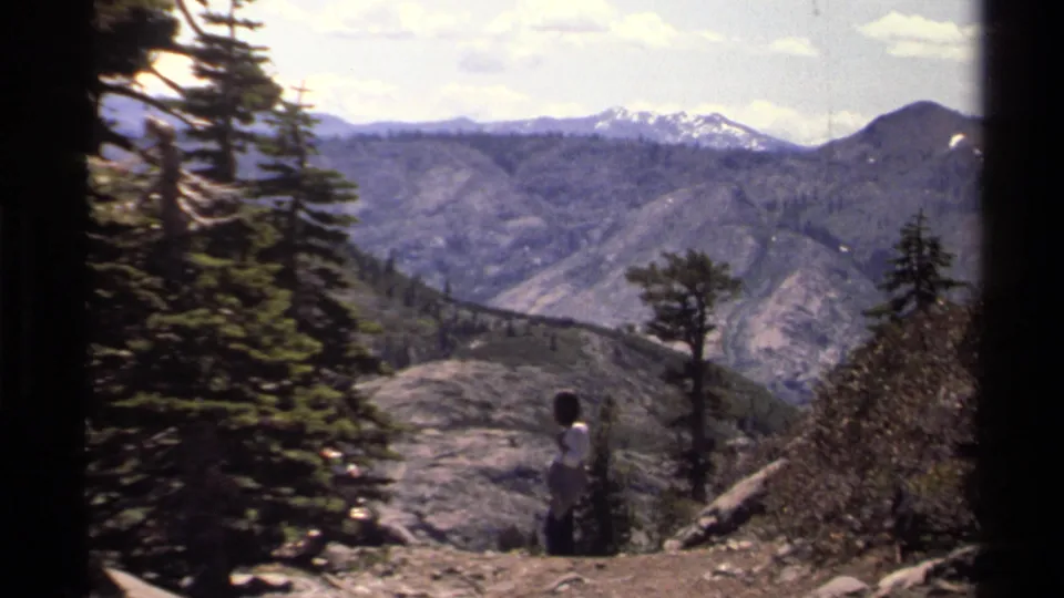 1978 Lake Tahoe home-movie frame of a woman walking on a rocky path bordered by pines and mountain views.