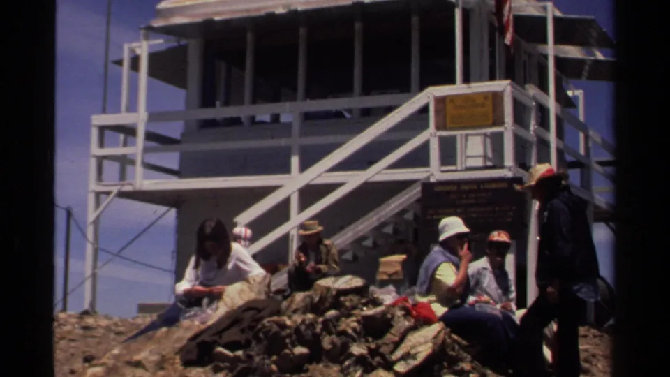 1978 Lake Tahoe home-movie frame showing visitors in hats and casual clothing resting near a white lookout tower and posted signs.