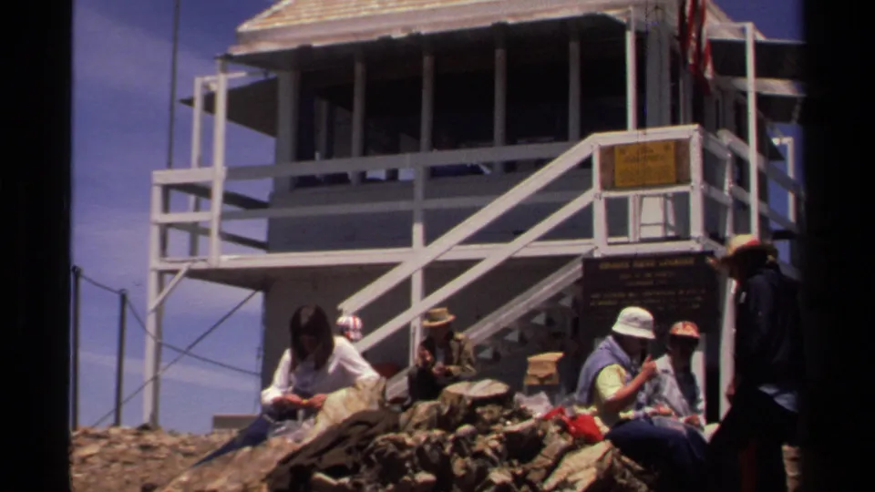1978 Lake Tahoe frame showing a small group beside a white lookout tower, with hats, bags, rocks, signs, and stairs visible.