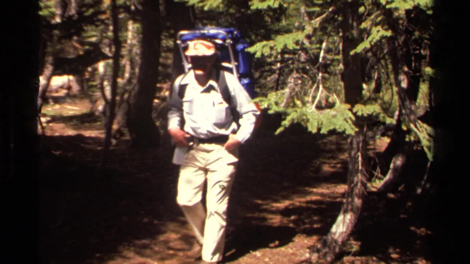 1978 Lake Tahoe home-movie frame showing a hiker with a blue backpack walking between pine trees on a forest trail.