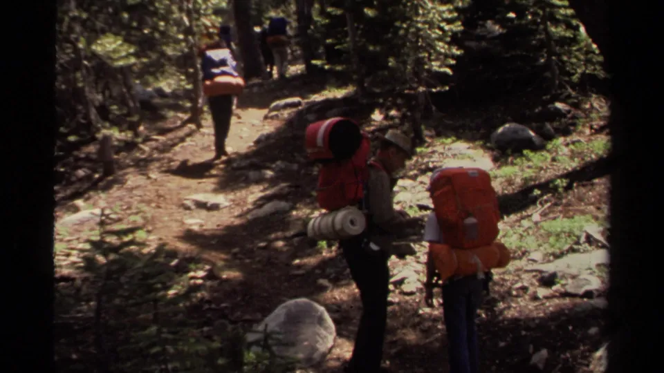 1978 Lake Tahoe frame with hikers spread along a shaded trail, including one person bent near a large orange backpack.