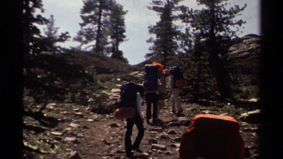 Lake Tahoe 1978 home-movie still with backpackers walking away on a shaded trail bordered by pines and rocks.