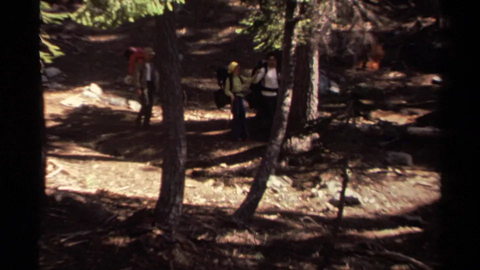 1978 Lake Tahoe home-movie frame showing several hikers with backpacks on a shaded forest trail among tall trees.