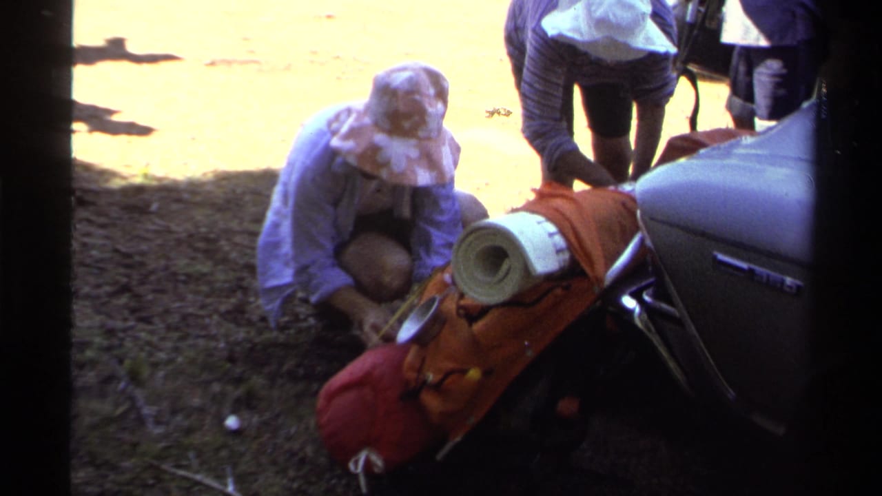 Person in a sun hat kneels near hiking gear beside a parked car.
