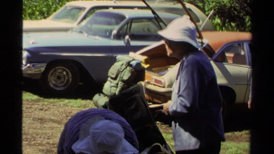 A hiker in a broad hat stands near external-frame backpacks and parked cars in the 1978 Lake Tahoe outdoor scene.