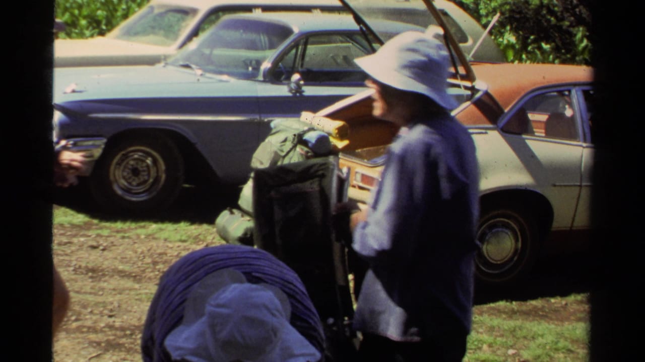 1978 Lake Tahoe home-movie frame showing adults preparing hiking gear beside parked cars in bright sun.