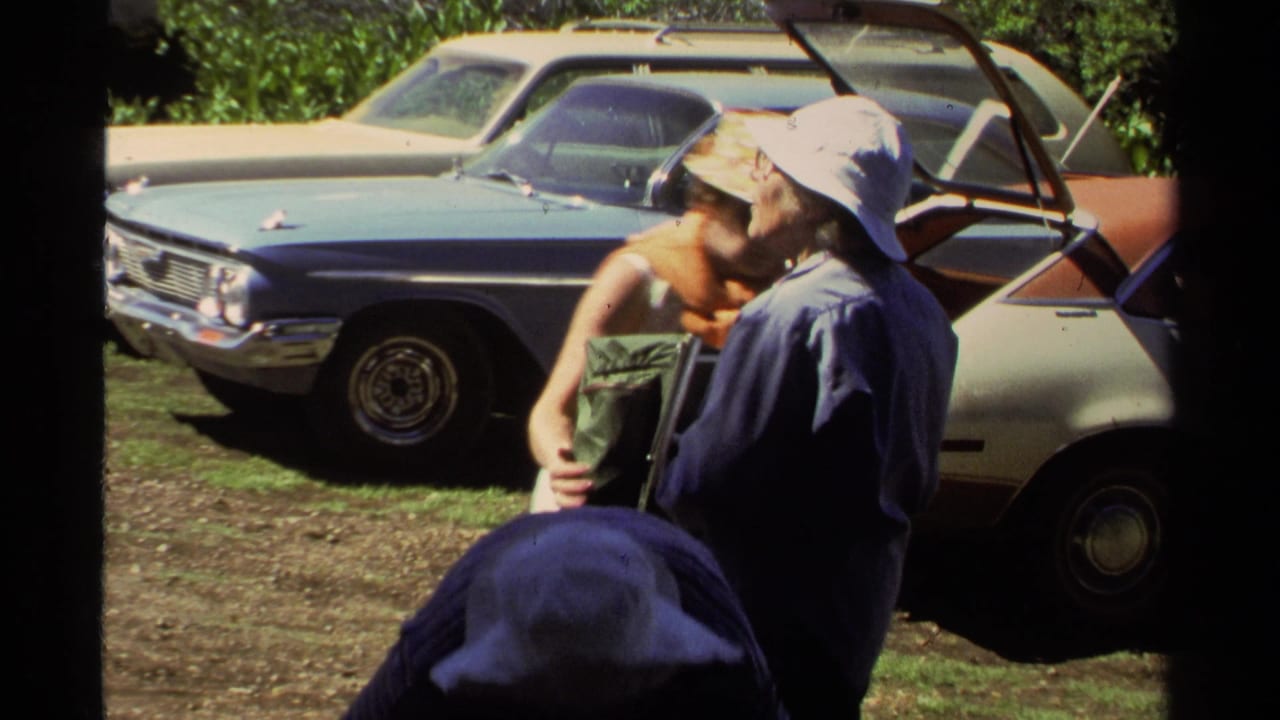 Two adults stand near an open car door while a backpack rests in front of them.