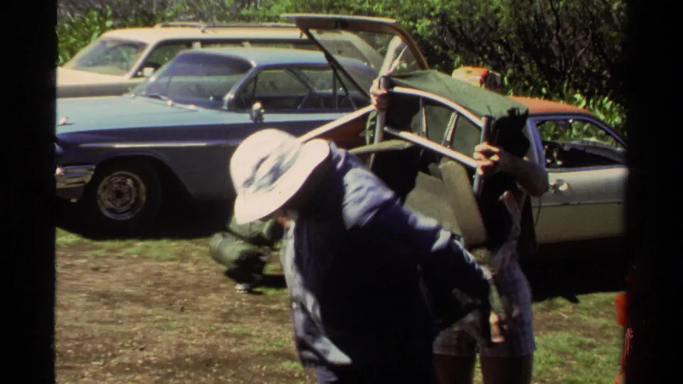 Home-movie frame from 1978 Lake Tahoe showing hikers adjusting a pack beside a car in a sunny lot.