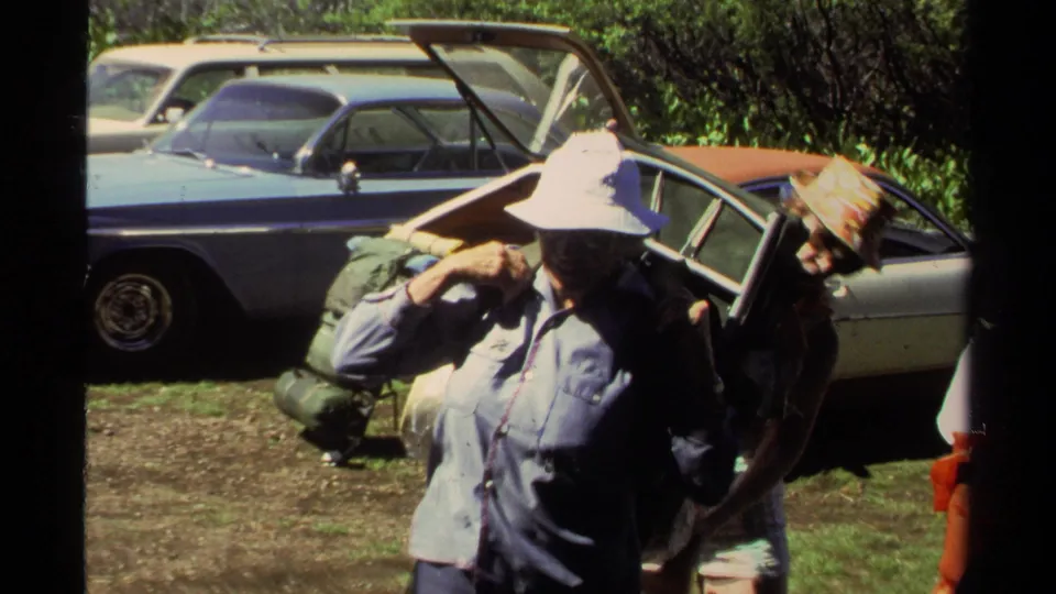 1978 Lake Tahoe frame showing an adult putting on a large backpack while another adult stands near the car.