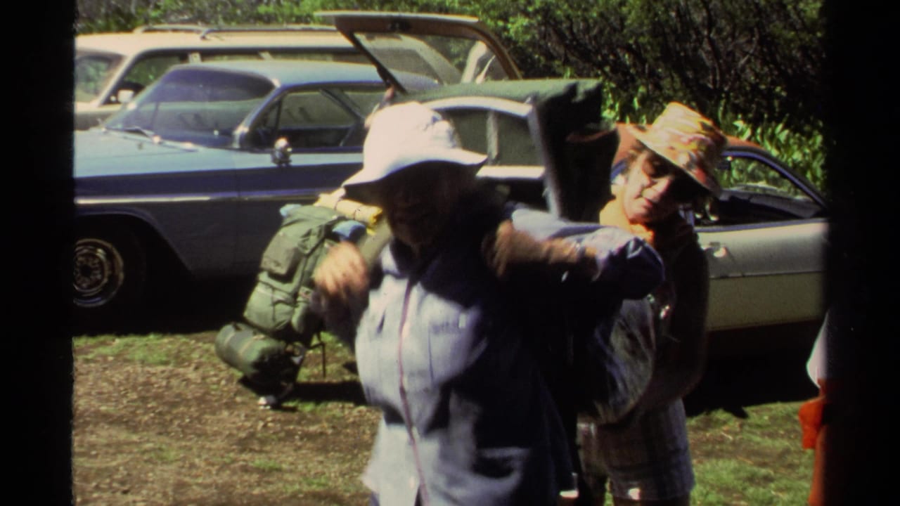 1978 Lake Tahoe home-movie frame showing adults adjusting outdoor packs beside parked cars in bright sunlight.