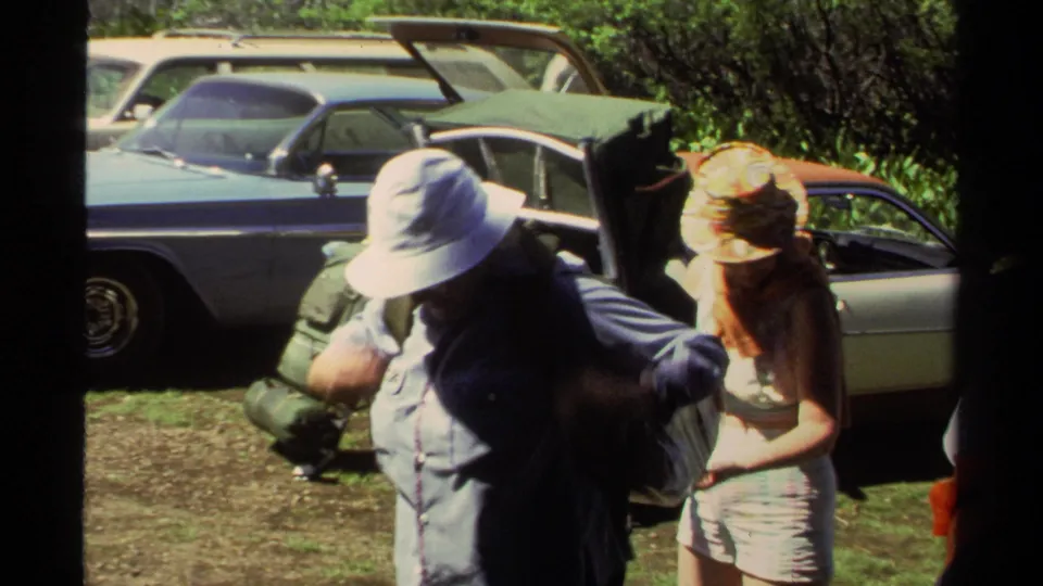 1978 Lake Tahoe home-movie frame with adults preparing bulky backpacks beside parked vehicles and trees.