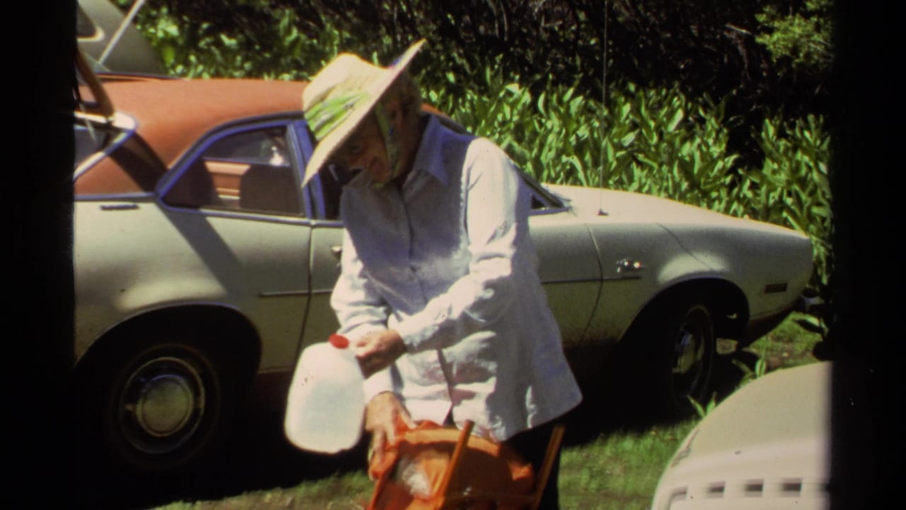 At Lake Tahoe in 1978, the woman bends toward an orange bag while holding a water jug beside a vintage car.