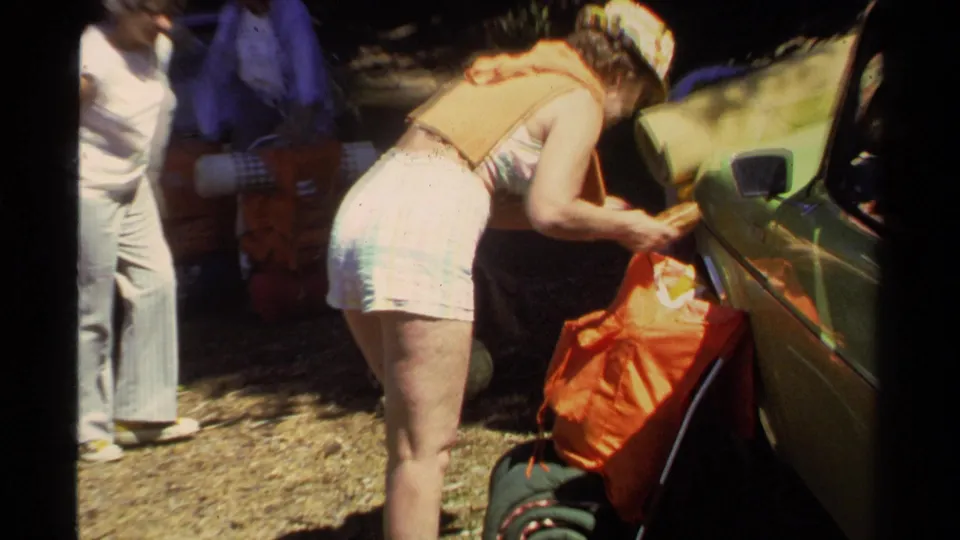 At Lake Tahoe in 1978, a person in a patterned skirt and orange vest packs supplies into an orange bag near a car.