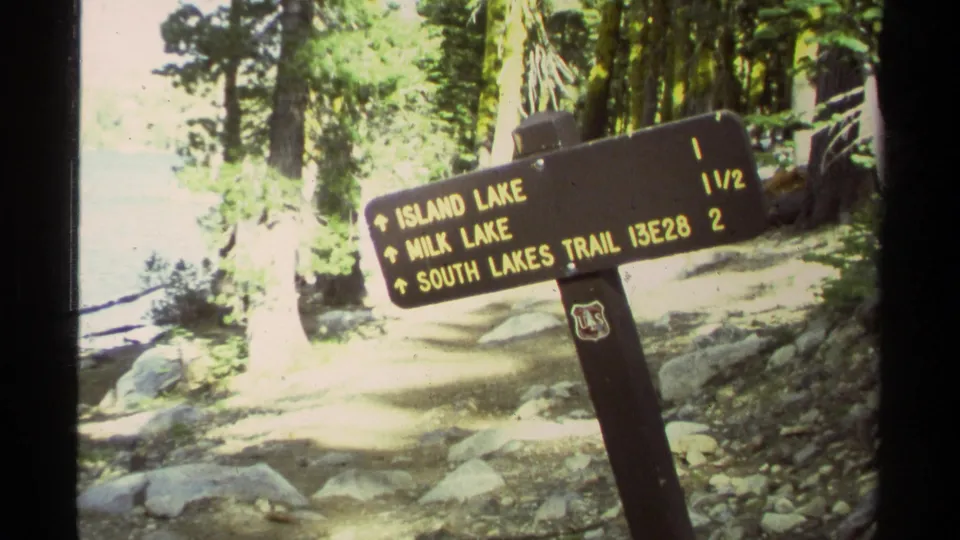 Forest trail sign lists Island Lake, Milk Lake, and South Lakes Trail, with trees, rocks, and lake-edge light behind it.
