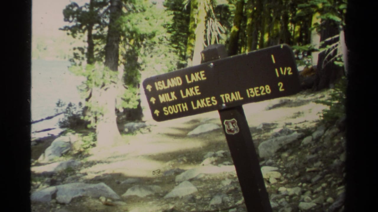 1978 Lake Tahoe-area home-movie frame showing a wooden trail sign for Island Lake, Milk Lake, and South Lakes Trail among trees and rocks.