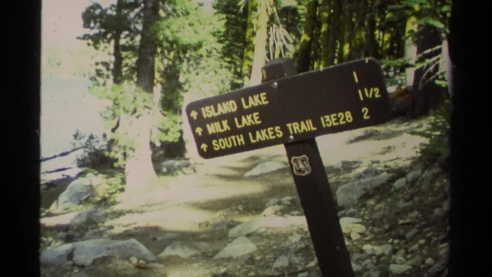 1978 Lake Tahoe-area home-movie frame showing a wooden trail sign for Island Lake, Milk Lake, and South Lakes Trail among trees and rocks.