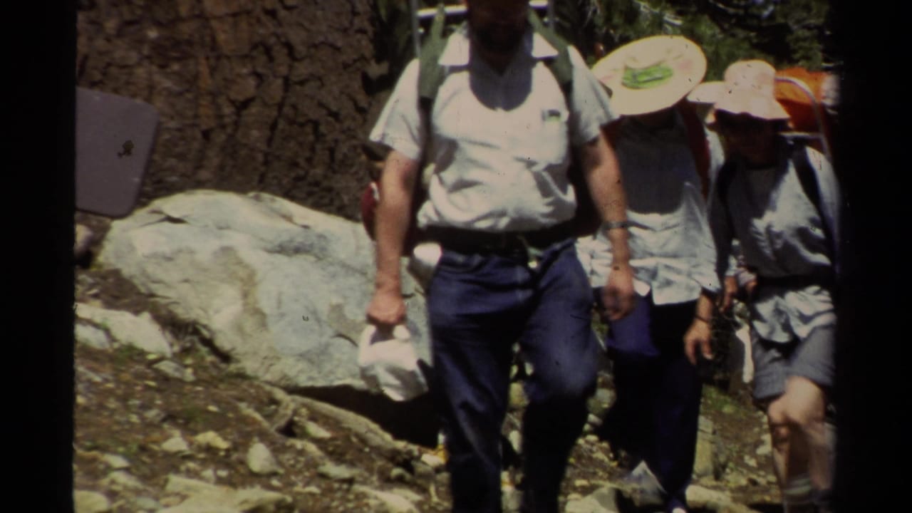 Family hikers in 1978 Lake Tahoe footage walk past boulders and pine trees, carrying backpacks and wearing sun hats on a mountain trail.