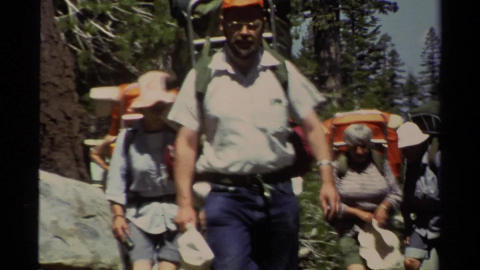 A close home-movie view shows several hikers with packs moving along a Lake Tahoe forest trail, including a man in a white shirt and orange-capped hiker.