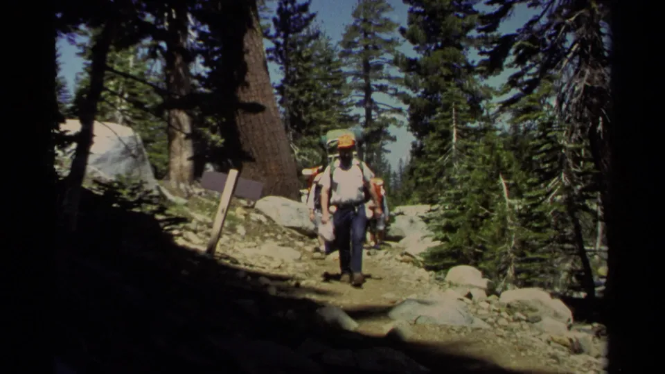 A wider 1978 Lake Tahoe hiking view shows a backpacked hiker in an orange cap walking through tall pines on a rocky mountain path.
