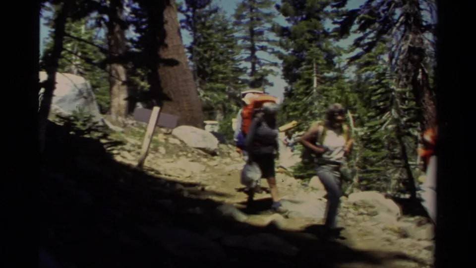 1978 Lake Tahoe 8mm frame of backpacked hikers continuing past the camera through a shaded conifer trail.