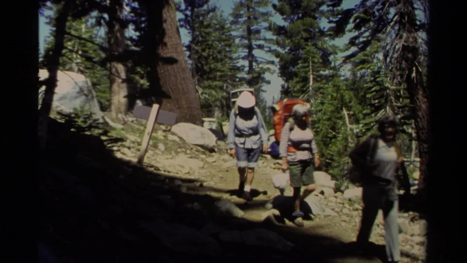 1978 Lake Tahoe home-movie frame showing hikers with large packs moving along a sunlit, rocky pine forest trail.