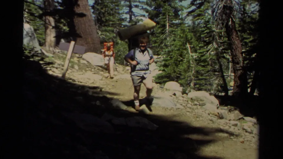 1978 Lake Tahoe home-movie frame of a hiker with a backpack and rolled pad moving through a pine forest trail.