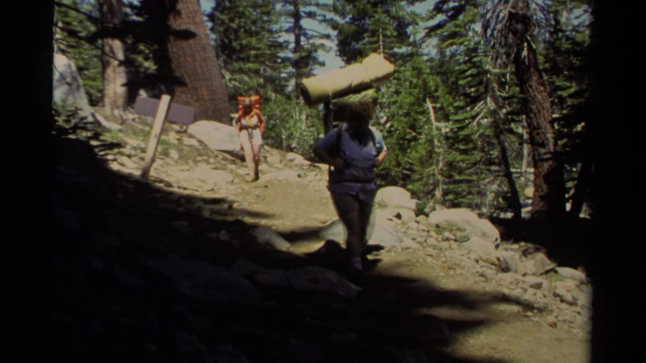 Two hikers on a rocky forest trail, one carrying a rolled sleeping pad and another following with a red pack.