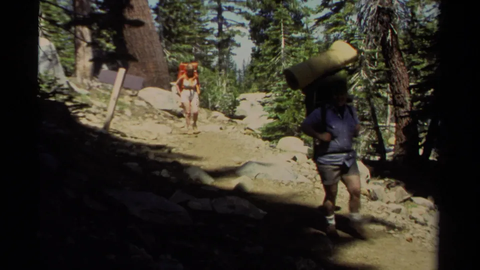 A hiker carrying a large rolled sleeping pad walks near the camera on a Lake Tahoe forest trail, with another backpacker behind.