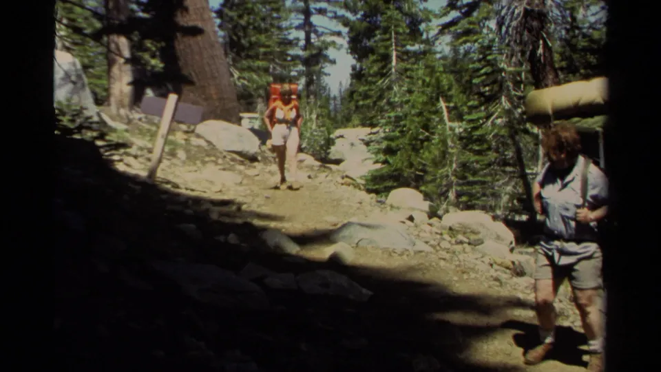 On a rocky Lake Tahoe trail, one adult hiker with a rolled pad stands near camera while another backpacker walks forward in the pines.