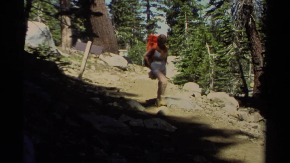 A backpacker with a red pack moves down a rocky Lake Tahoe forest trail in 1978 8mm home-movie footage.