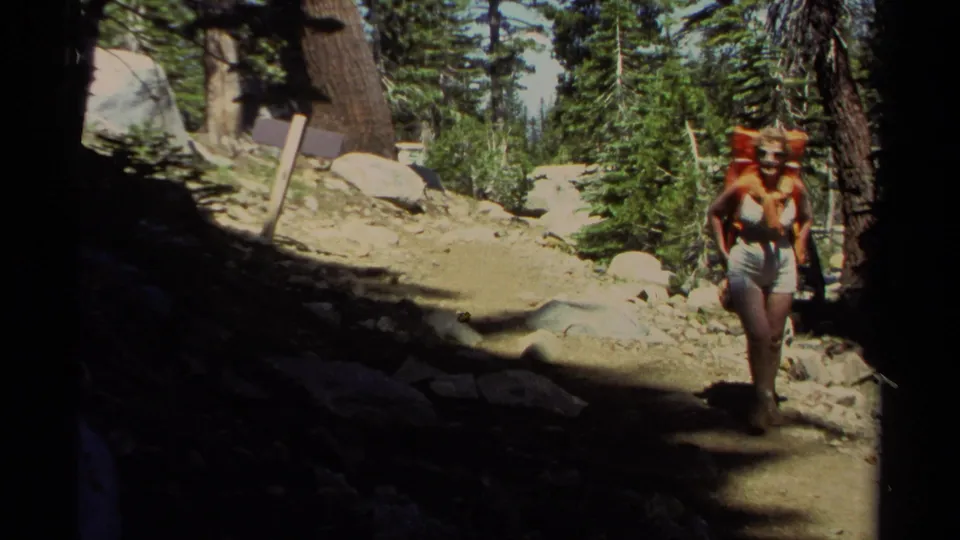 A woman in shorts and a light top hikes toward the camera on a rocky Lake Tahoe forest trail, carrying a large orange pack.