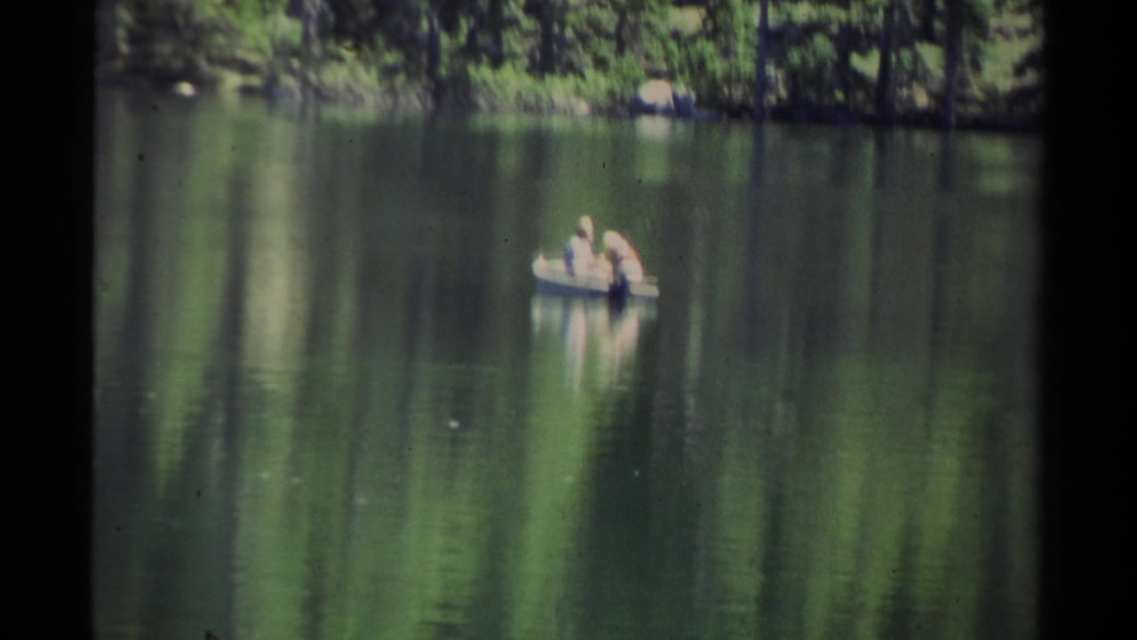 Two people sit in a small boat on calm lake water near a wooded shoreline.