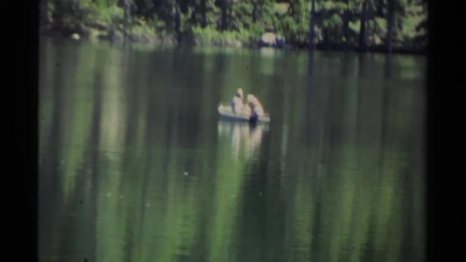 1978 Lake Tahoe home-movie frame showing two distant people in a small boat on reflective water with evergreen trees behind them.