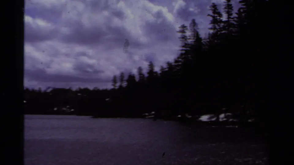 1978 Lake Tahoe home-movie view of a dark evergreen shoreline beneath a cloudy sky, with lake water in the foreground.