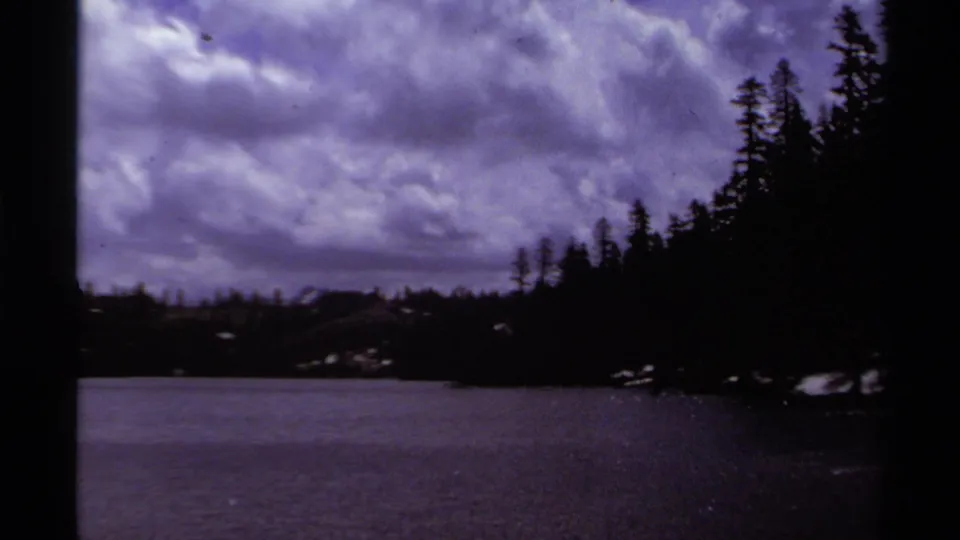 A late-1970s Lake Tahoe frame shows a shadowed evergreen shore, open lake water, and a purple-toned cloudy sky on 8mm film.