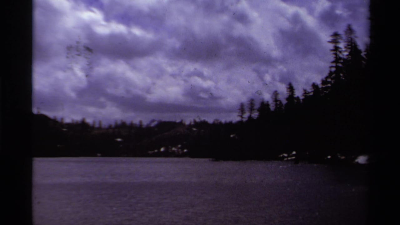 1978 8mm Lake Tahoe view with calm water, dark pines, and purple-blue clouds across the sky.