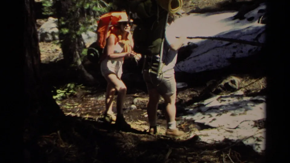 Two adult backpackers in 1978 Lake Tahoe footage stand close on a wooded trail with external-frame packs and snow behind them.