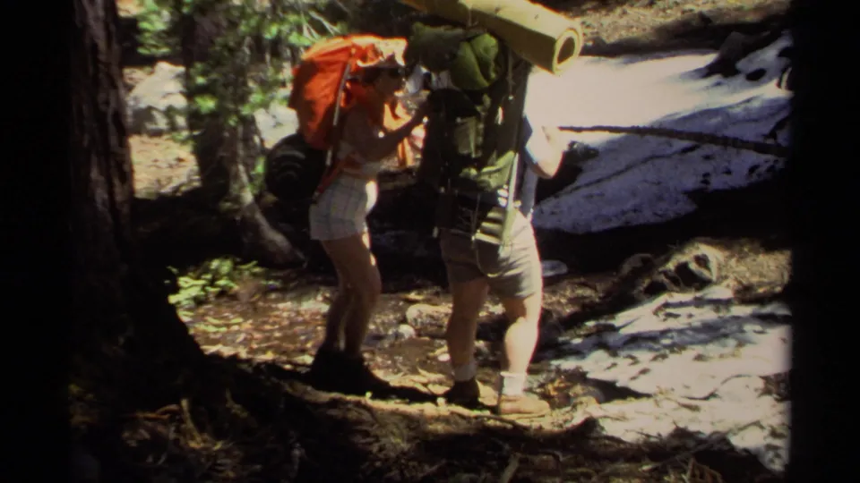 A pair of adult hikers with large packs pause together on a shaded Lake Tahoe forest trail, with bright snow visible behind them.