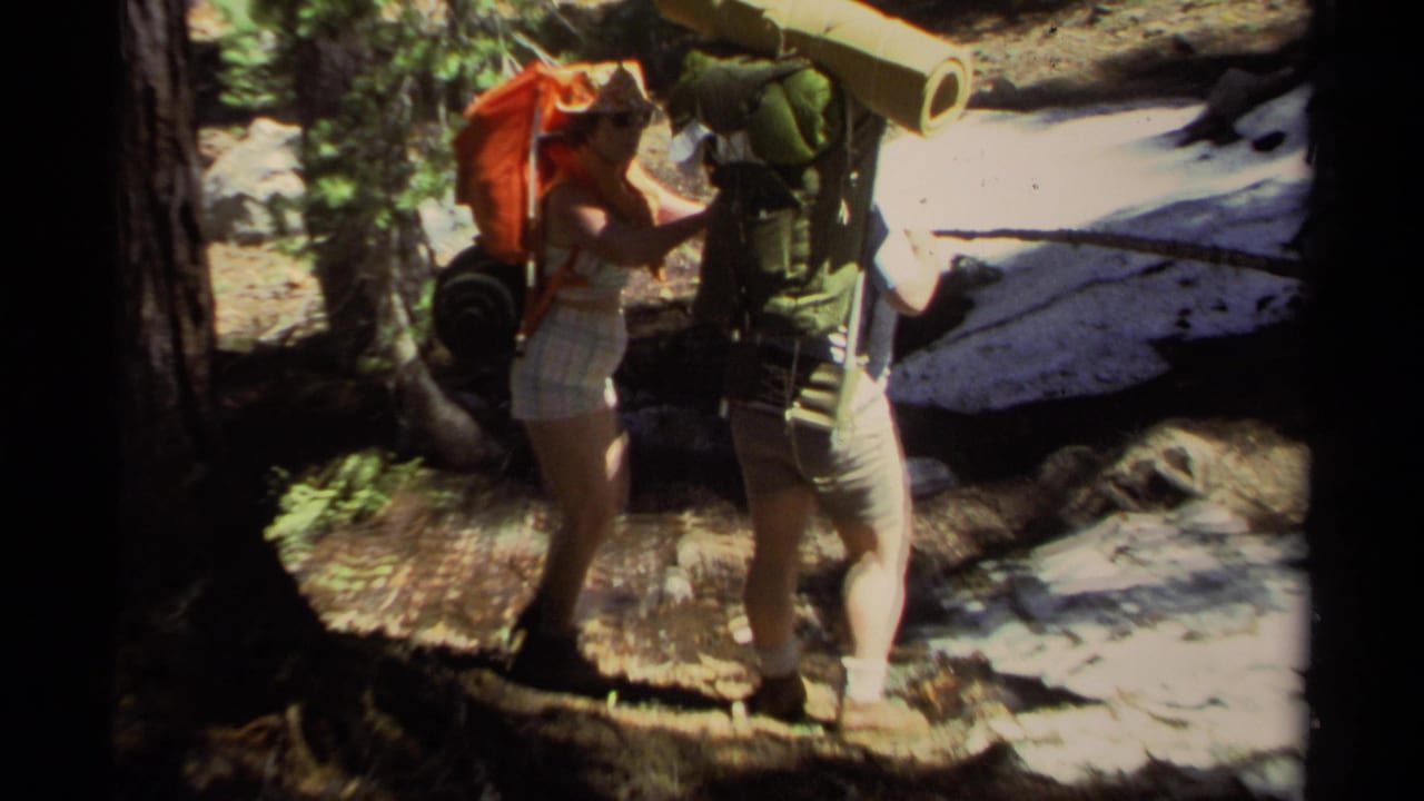 Two hikers near Lake Tahoe in 1978 pause on a wooded trail with large packs, a rolled mat, and dappled forest light.