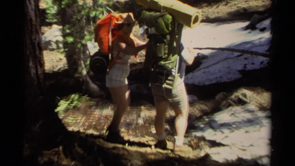 Two hikers near Lake Tahoe in 1978 pause on a wooded trail with large packs, a rolled mat, and dappled forest light.