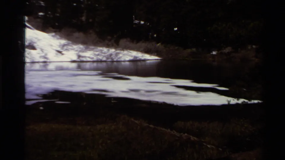 1978 Lake Tahoe footage frame showing dark water in the foreground and melting snow along a shaded evergreen shoreline.