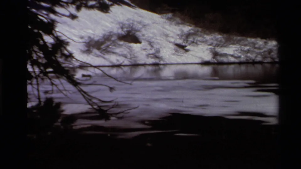 1978 Lake Tahoe home-movie frame showing a snowy shoreline across still water, partially framed by dark branches and tree shadow.