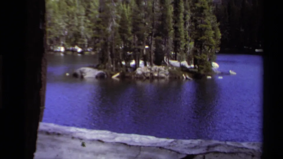 Lake Tahoe 8mm frame showing a small evergreen island, rocks, blue water, and patches of lingering snow.