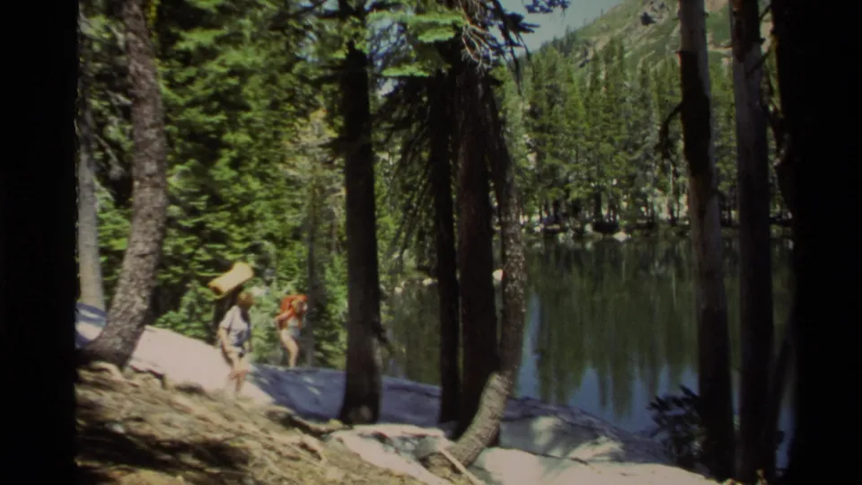 In Lake Tahoe home-movie footage from 1978, two backpacked hikers move through pine trees near a reflective lakeshore.