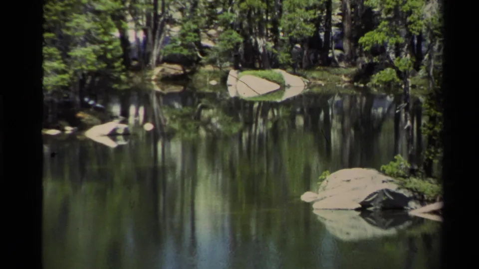 A quiet Lake Tahoe shoreline view shows still water, granite boulders, and dense evergreens reflected on the lake surface.